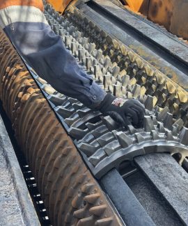 A worker wearing gloves adjusts or inspects large interlocking metal rollers with sharp, gear-like teeth inside an industrial machine, likely used for crushing or processing materials.