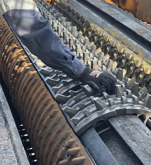 A worker wearing gloves adjusts or inspects large interlocking metal rollers with sharp, gear-like teeth inside an industrial machine, likely used for crushing or processing materials.