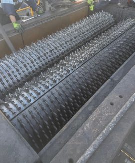 Top view of an industrial machine with multiple large rotating rollers covered in sharp, interlocking teeth, with workers wearing gloves positioned around the equipment for maintenance or inspection.